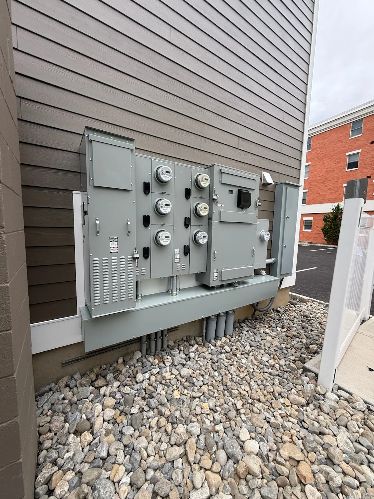 Gray steel electrical panel with six meters and disconnect switches on gravel pad against building