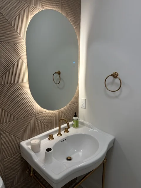 Illuminated oval mirror with LED backlight above white pedestal sink with brass fixtures