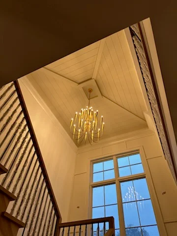 Gold chandelier installed in vaulted foyer with geometric ceiling details and tall windows